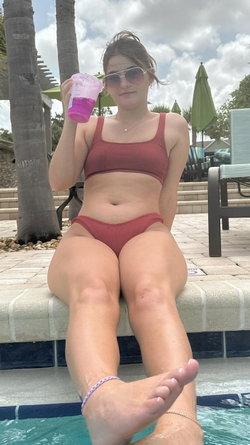Curvy woman in red bikini sits poolside holding a drink wearing sunglasses with palm trees behind.
