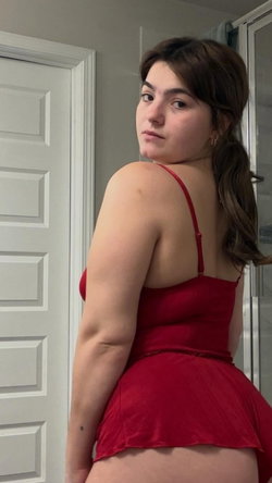 Curvy brunette in a red dress looks back over her shoulder in an indoor setting.