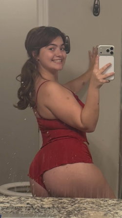 Curvy brunette in a red dress taking a selfie in a bathroom shower.