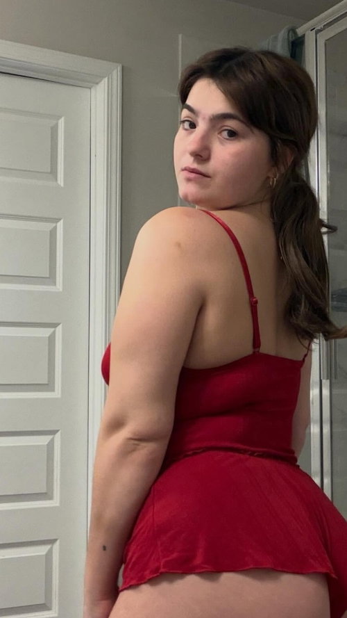 Curvy brunette in a red satin dress poses in a bathroom, glancing back over her shoulder.