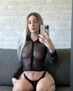 A woman with long blonde hair sits on a gray couch, wearing a sheer black button-up shirt and black lace underwear,