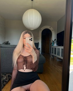 Blonde woman in sheer brown top and black skirt takes mirror selfie in living room with wooden floors and modern lamp.