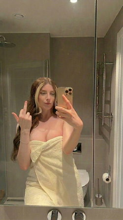 A woman with long brown hair and blonde highlights takes a mirror selfie in a bathroom, wrapped in a yellow towel,