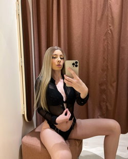 Blonde woman in sheer black shirt and lace underwear poses on a stool, taking a mirror selfie in a room with brown