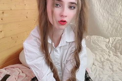 A young woman with long wavy hair and red lipstick sits on a bed wearing an unbuttoned white shirt, looking directly at