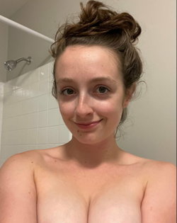 A woman with brown hair in a messy bun smiles softly in a bathroom, bare-chested with shoulders and upper chest visible.