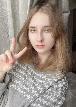 A young woman with light brown hair and green eyes makes a peace sign while wearing a gray patterned sweater indoors.