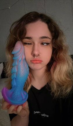 A young woman with wavy blonde hair and a nose ring holds a colorful toy close to her face, wearing a black shirt in a