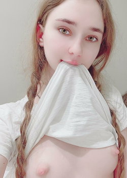 A young woman with long light brown hair and fair skin bites a white fabric, revealing bare breasts while wearing a