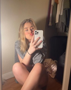 Blonde woman in a gray shirt takes a mirror selfie, sitting on the floor with legs bent, making a playful face.