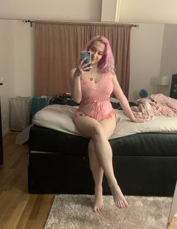 A person with pink hair in a pink nightie sits on a bed taking a selfie.