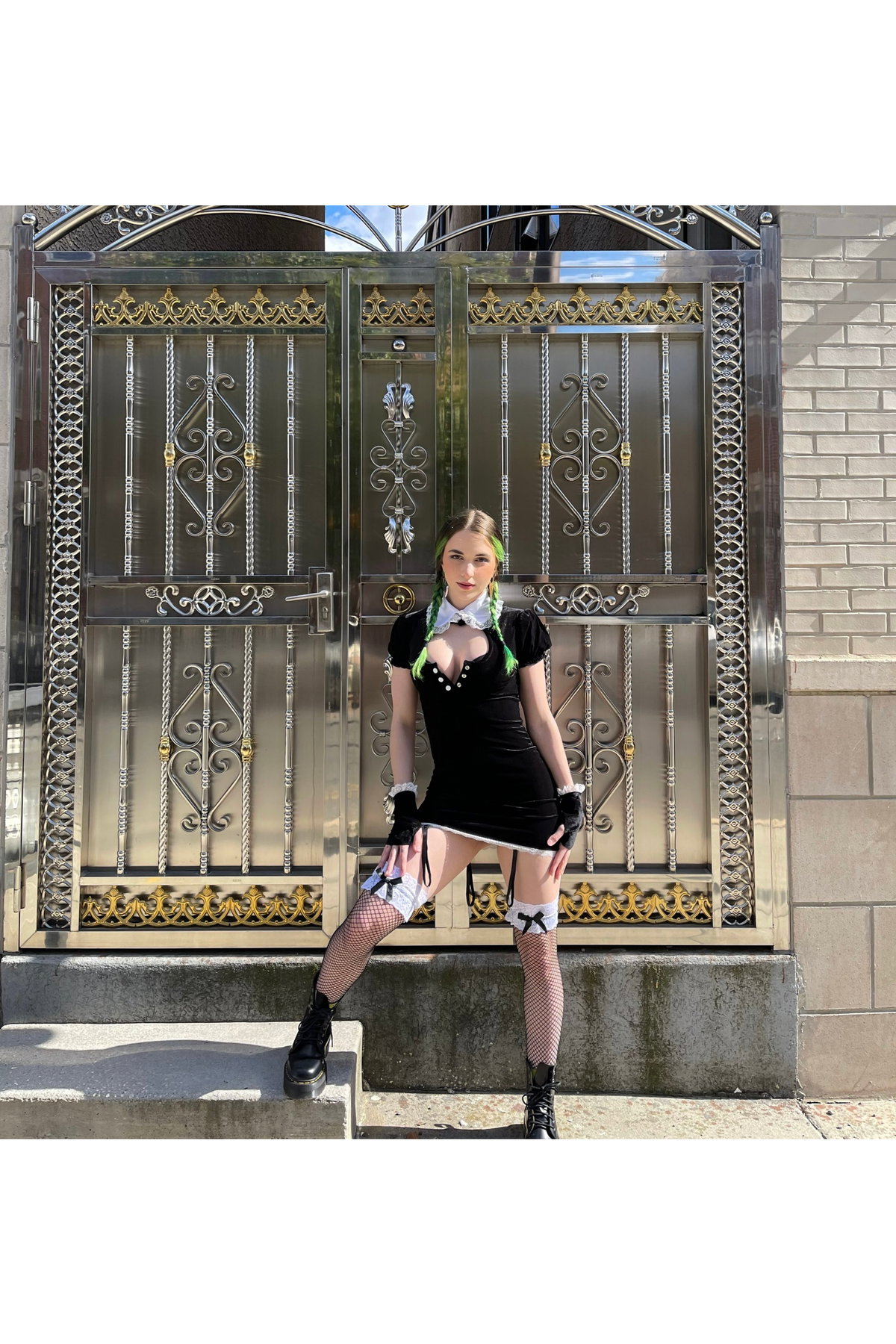 A woman with green-tipped braids wears a black dress with a white collar, fishnet stockings, and black boots, posing in