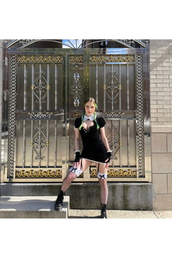 A woman with green-tipped braids wears a black dress with a white collar, fishnet stockings, and black boots, posing in