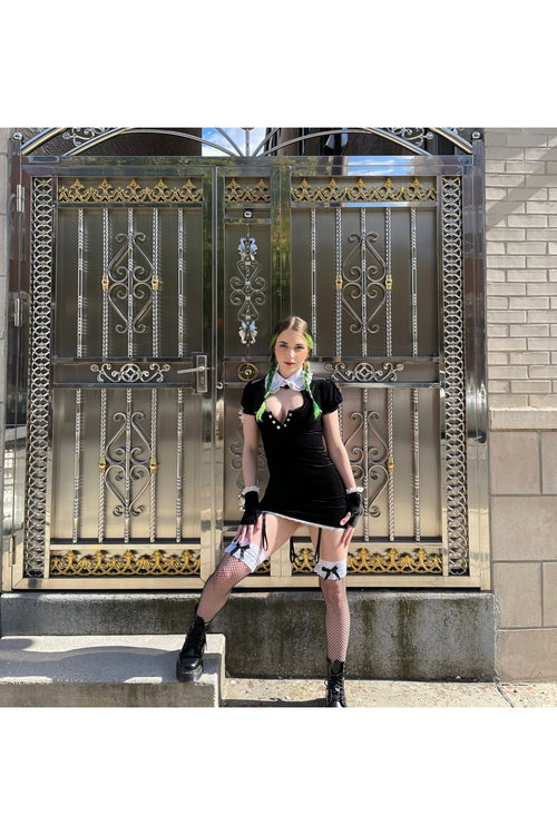 A woman with green-tipped braids wears a black dress with a white collar, fishnet stockings, and black boots, posing in