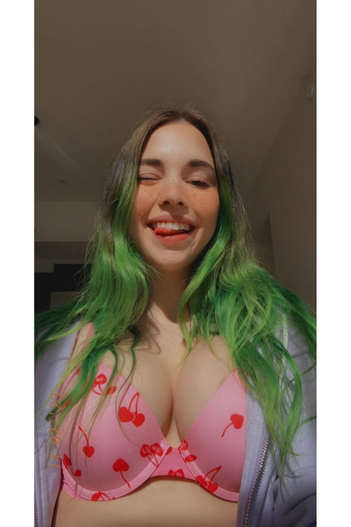 A woman with green-tipped hair smiles in a pink cherry-print bra and light jacket, indoors.