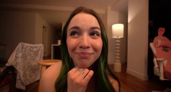 A young woman with green-tipped hair smiles playfully in a cozy indoor setting, hand resting under her chin.