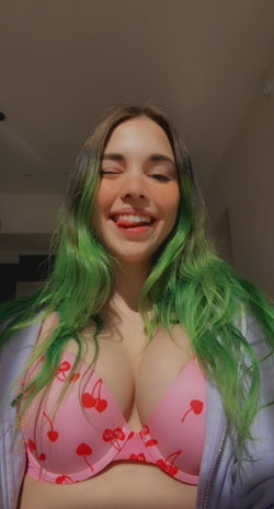 A smiling woman with green-tipped hair wears a pink cherry-print bra and a light jacket, taking a selfie indoors.