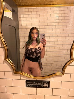 A young woman with green-tipped hair takes a mirror selfie in a tiled bathroom, wearing a floral crop top and showing