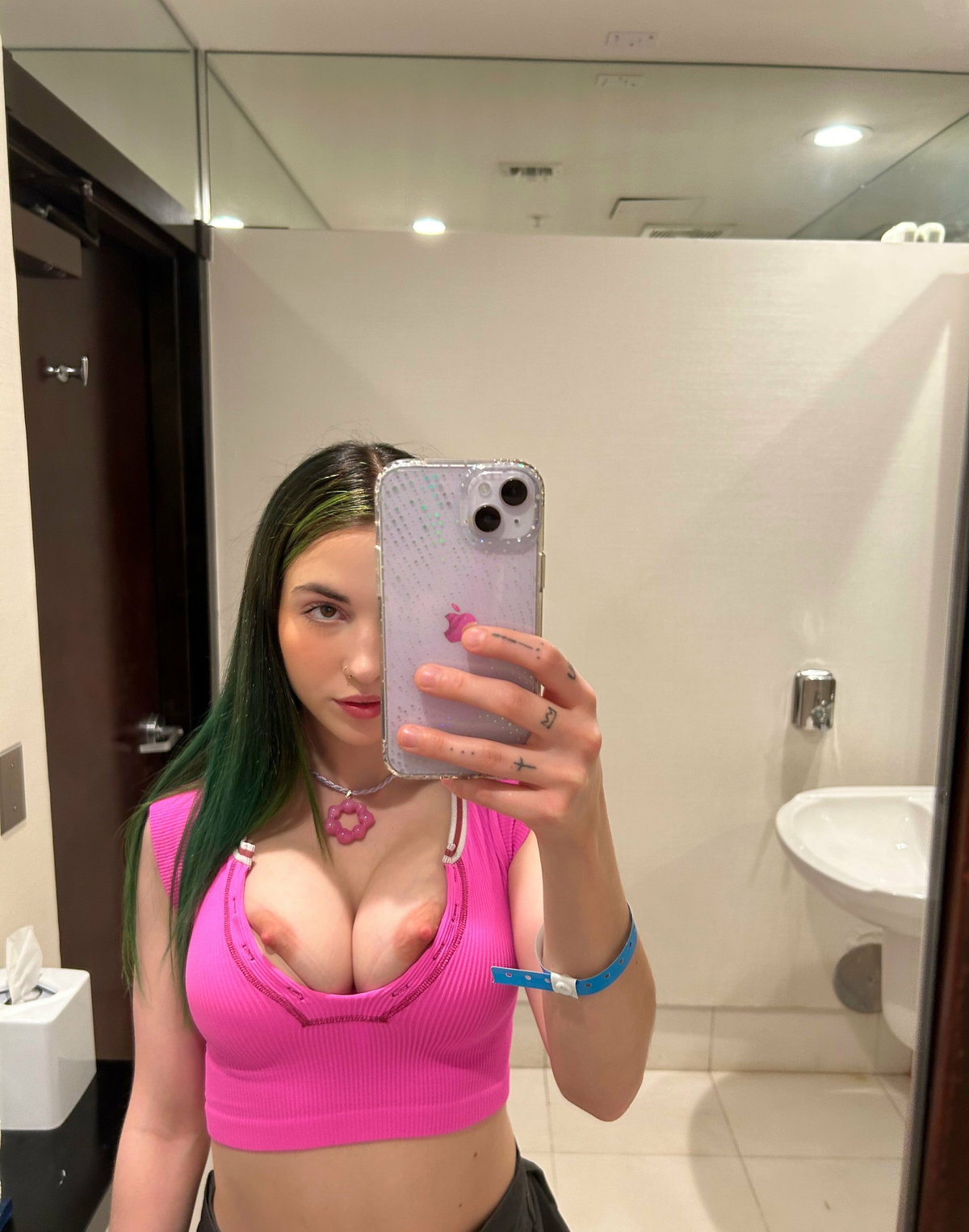 Woman with green hair takes mirror selfie in bathroom, wearing a pink crop top with visible nipples, tattoos on