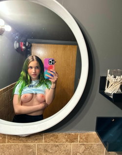 Woman with green-tipped hair takes mirror selfie in bathroom, wearing a knotted crop top and black bottoms, bare