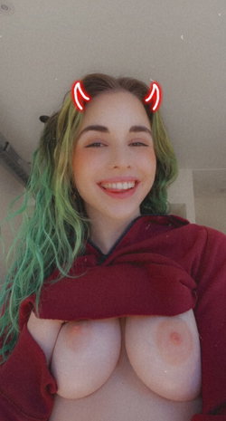 Woman with green hair and red devil horns filter, smiling while holding open a red hoodie to expose her bare breasts.