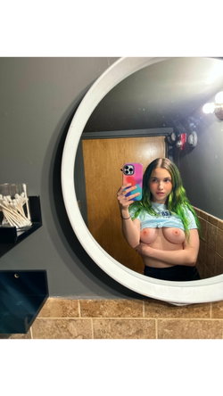 A young woman with green-tipped hair takes a mirror selfie in a bathroom, wearing a knotted crop top that exposes her