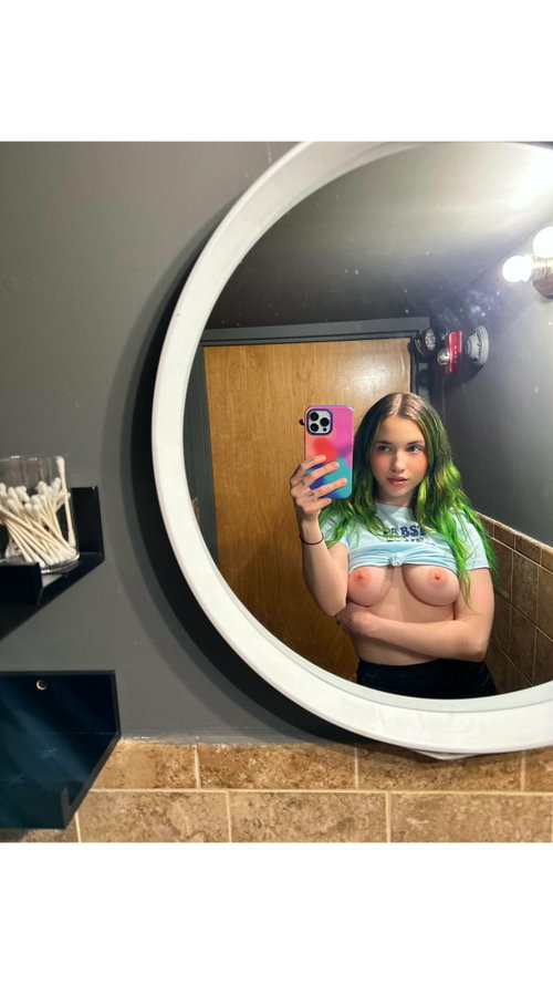 A young woman with green-tipped hair takes a mirror selfie in a bathroom, wearing a knotted crop top that exposes her