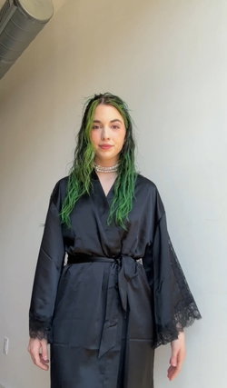 A person with green hair and a black robe stands indoors.