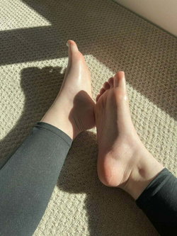 Close-up of bare feet with painted toenails, wearing dark leggings, resting on a carpeted floor in sunlight.