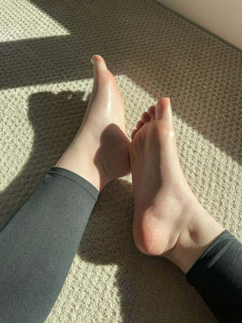 Close-up of bare feet with painted toenails, wearing dark leggings, resting on a carpeted floor in sunlight.
