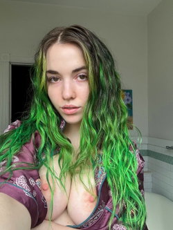 A woman with long green hair and a nose ring takes a selfie in a bathroom, wearing an open purple robe with bare