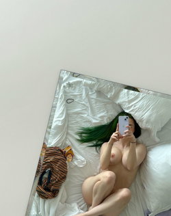 A woman with vibrant green hair lies nude on a white bed, taking a mirror selfie with her phone, surrounded by