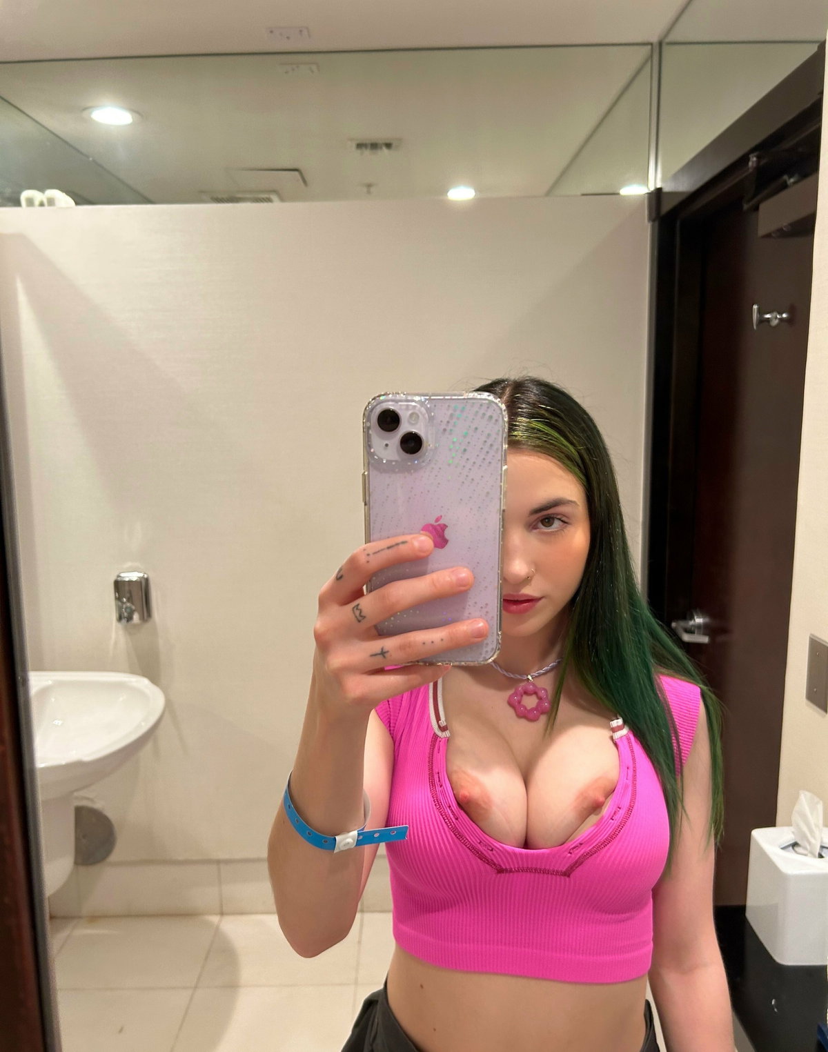 A woman with green-tipped hair takes a mirror selfie in a bathroom, wearing a tight pink crop top that exposes her