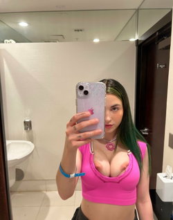 A woman with green-tipped hair takes a mirror selfie in a bathroom, wearing a tight pink crop top that exposes her