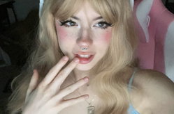 Blonde woman with bangs, heavy makeup, nose ring, and white cat ears poses closely with hand near lips, wearing a light