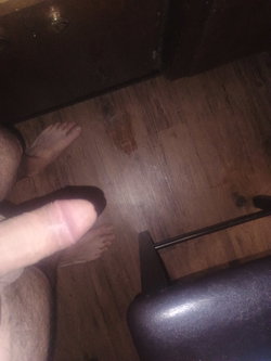 Close-up of bare feet on wooden floor, partial leg visible, taken from above in dimly lit room with furniture nearby.