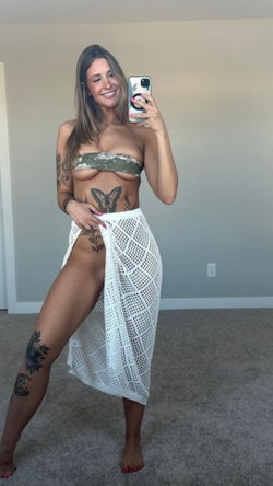 A smiling tattooed brunette takes a selfie indoors while wearing an open white skirt.