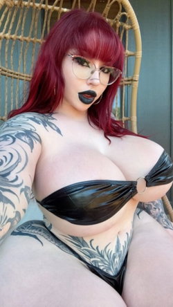 Curvy redhead with glasses and tattoos sits in a wicker chair wearing black swimwear.