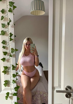 Blonde woman in pink lingerie takes mirror selfie in bedroom with green vines and striped lamp.
