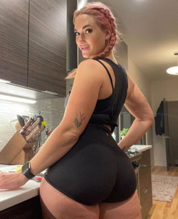A woman with pink braided hair in a black athletic outfit poses in a kitchen, showing a curvy figure with visible
