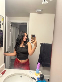 Brunette woman in black crop top and red skirt takes mirror selfie in bathroom, showing midriff and navel piercing.