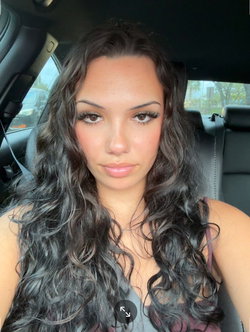 Brunette woman with long wavy hair and subtle makeup, wearing a dark tank top, sitting in a car interior.
