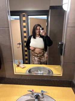 A woman with dark hair and glasses takes a mirror selfie in a bathroom, wearing a white top, black jacket, and