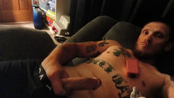 A tattooed man with a beard lies shirtless on a couch, holding his penis, with multiple visible tattoos on his chest