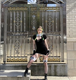 A woman with green-tipped braids wears a black dress with white lace collar, fishnet stockings, and garters, posing in