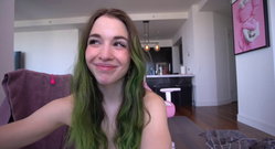 A woman with long green-tipped hair smiles playfully in a modern living room, wearing no top, with a pink artwork