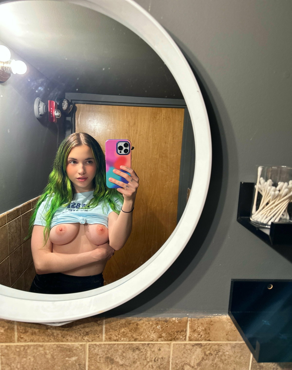 A woman with green-tipped hair takes a mirror selfie in a bathroom, wearing a knotted crop top and black bottoms, bare