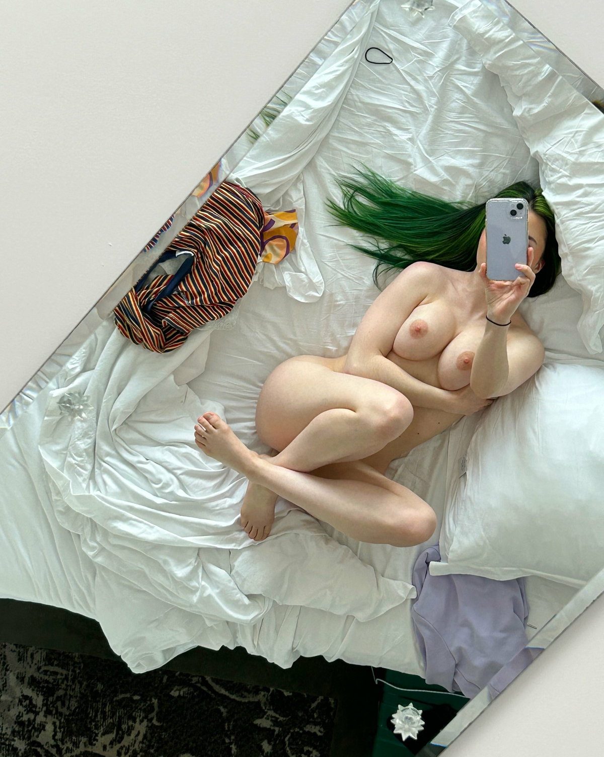A person with vibrant green hair lies nude on a white bed, taking a selfie with a phone, arms covering breasts, legs