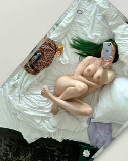 A person with vibrant green hair lies nude on a white bed, taking a selfie with a phone, arms covering breasts, legs