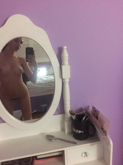 A woman with a curvy build takes a mirror selfie in a bedroom with purple walls, showing her bare torso while holding a